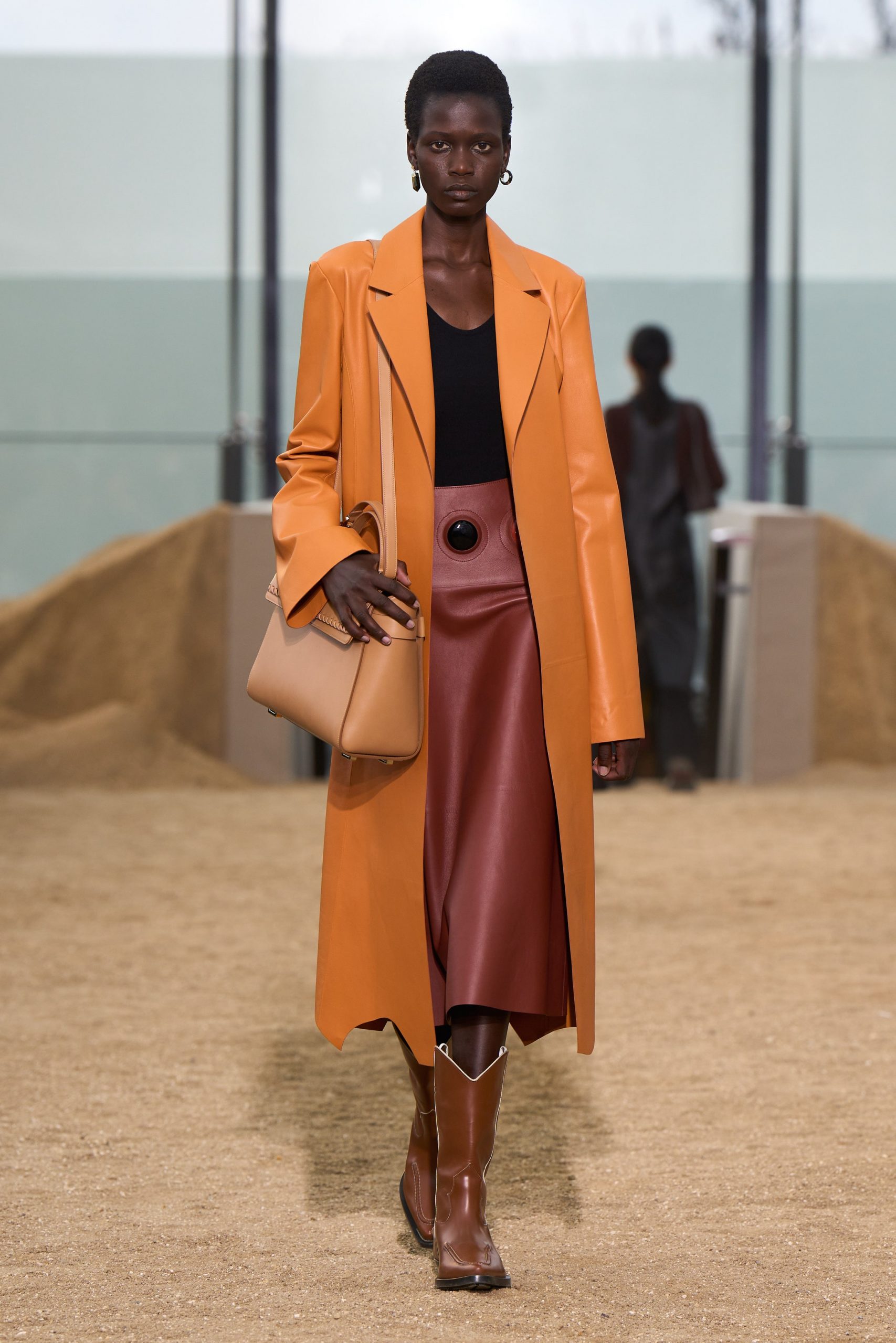 A model wears a black top, leather knee-length skirt, cowboy boots, and a leather coat