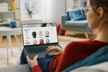 Young Woman at Home Using Laptop Computer for Browsing Through Online Retail Shopping Site. She's Sitting On a Couch in His Cozy Living Room. Over the Shoulder Camera Shot
