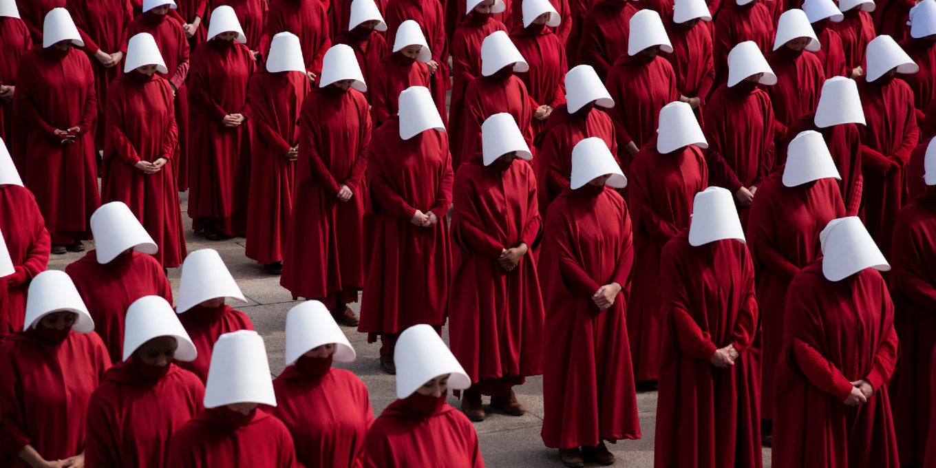 A series of women stand in lines wearing red dresses and white bonnets