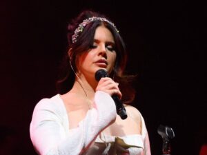 Lana Del Rey performing at Glastonbury music festival in a white long-sleeve square neck dress with diamond headband and her hair up in a loose bun with face framing bangs.