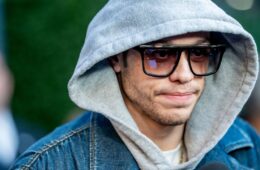 Pete Davidson wearing a jean jacket with a grey hoodie underneath pulled up on his head and black sunglasses on his face.