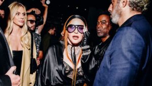 Madonna partying in a black lace top and black satin jacket. she has red hair pushed back by a black satin headband and  is wearing black sunglasses with purple lenses