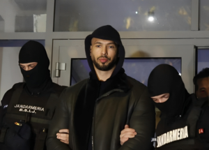Andrew Tate, in a black leather jacket and a black hoodie with the hood pulled up being escorted by Romanian police who are in black ski masks and bulletproof vests reading "JANDARMERIA B.S.I.J"