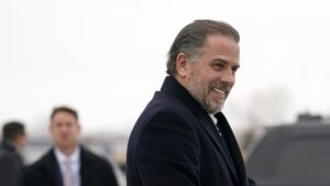 Hunter Biden smiling off to the right in a blue coat and white undershirt. There are two people off to his left in the blurred background.