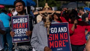 Two Catholics for Catholics protestors. The protestor on the right holding a large crucifix and a sign that says "go and sin no more John 8:11" and on the left holding a sign that says "sacred heart of Jesus have mercy on us"