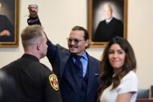 Johnny Depp smiling and raising their fist in the courtroom. To his front right is his lawyer Camille Vasquez grinning to the side in a white dress. To Depp's front left is a police officer facing him.