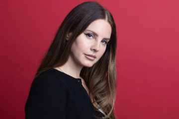 Lana Del Rey posing in front of a deep red background, with the body facing to the right and looking at the camera. she has her medium brown hair, with highlights, down and slightly curled, and is wearing a black long sleeve shirt.