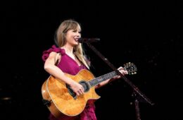 Taylor Swift performing the 'speak now' section at the 'eras tour'. she's in front of a mic stand wearing a maroon dress with her hair down and she's holding a light brown acoustic guitar.
