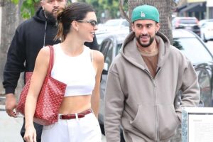 Kendall Jenner and Bad Bunny walking together on a shopping date. Kendall is in a white tank top and white pants with a burgundy belt and a red handbag on her shoulder. Bad bunny is wearing a grey hoodie with a medium brown shirt underneath and a light green hat