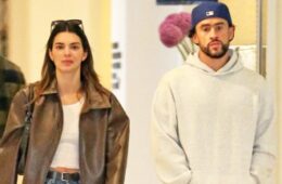 Kendall Jenner and Bad Bunny walking together. Bad Bunny is in a grey sweatshirt and a blue hat. Kendall is wearing a white crop top with a large light brown leather jacket, blue jeans with a black belt and a gold clasp, black sunglasses on her hair and a black handbag on her shoulder.