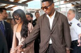 Jonathan Majors, in a light brown suit with white shirt, walking hand in hand with girlfriend Meagan Good, in a pink suit jacket and white low-cut shirt. a crowd is being them along with police officers preventing people from approaching them.