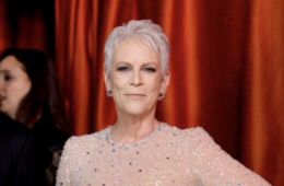 Jamie Lee Curtis posing for a photo at the oscars. She is in front of a red velvet curtain in a light-pink/cream sequined dress