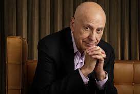 Alan Arkin posing for a photo. He is pictured seated with his hands entwined under his chin.