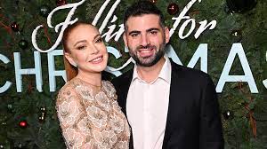 Lindsay Lohan with her husband Bader Shammas posing on the red carpet for her nettles movie 'Falling for Christmas'. Lindsay is in a cream dress with white flowers embroidered and Bader is in a black suit jacket with a white collard shirt.