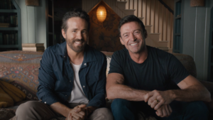 Ryan Reynolds (left) and Hugh Jackman (right) smiling on a brown couch together promoting Deadpool 3