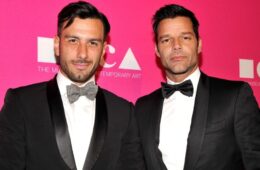 Ricky Martin and Jwan Yosef posing in front of a hot pink background wearing tuxedos. Jwan is wearing a grey bow tie and Ricky is wearing a black bow tie