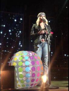 Miranda Lambert singing at a mic stand with a fan's beachball to the left on stage with her.