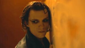 A screen shot of Tom Holland in 'The Crowded Room'. He is leaning forward against a wooden surface in black winged eye makeup.