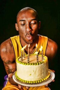 Kobe Bryant sitting in the dark, in his Lakers jersey, holding a birthday cake and blowing out the candles.