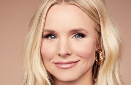 A headshot of Kristen Bell grinning in front if an ombre peach/light pink background.