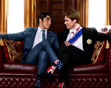 Alex Claremont-Diaz (left) sitting next to Prince Henry (right) on a leather couch with their legs crossed, smiling at each other