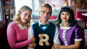 Betty (left) and Veronica (right) holding onto Archie's arms (middle) all sitting in a booth at 'POP's