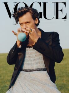 Harry Styles on the cover of Vogue Magazine wearing a baby blue lace dress with black trimming and a black blazer on top. He's blowing into a small baby blue balloon and is set on a field of green grass.