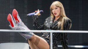Taylor Swift performing 'The Man' at the Eras Tour. She's in a black rhinestone stripped suit jacket with her silver, red bottomed, boots crossed up on a rail.