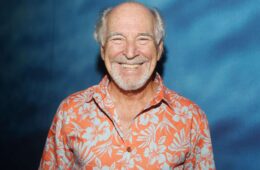 Jimmy Buffett smiling for a photo in an orange Hawaiian shirt standing in front of a blue background