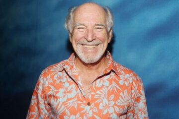 Jimmy Buffett smiling for a photo in an orange Hawaiian shirt standing in front of a blue background