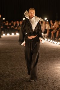 Model wearing a sculptural black Schiaparelli SS26 suit with an exaggerated white ruffled collar and wide-leg trousers walking the circular runway.