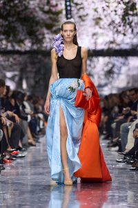 Model walks Dior Couture runway in light blue long skirt with bright orange purse and sheer black tank top 