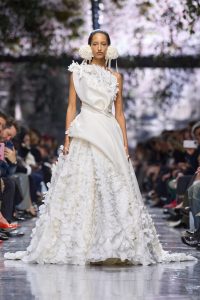 Model Walks Runway in Dior Couture show in white gown with draped bodice and floral skirt.