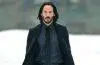 Keanu Reeves smiles during a public appearance, wearing a dark suit and his signature long hair and beard.
