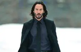 Keanu Reeves smiles during a public appearance, wearing a dark suit and his signature long hair and beard.