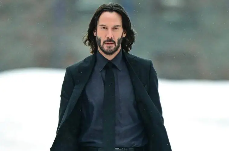 Keanu Reeves smiles during a public appearance, wearing a dark suit and his signature long hair and beard.