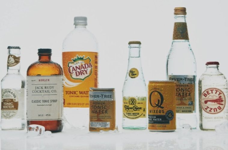 A chilled glass of gin and tonic with tonic water bottles and citrus garnishes arranged on a home bar.