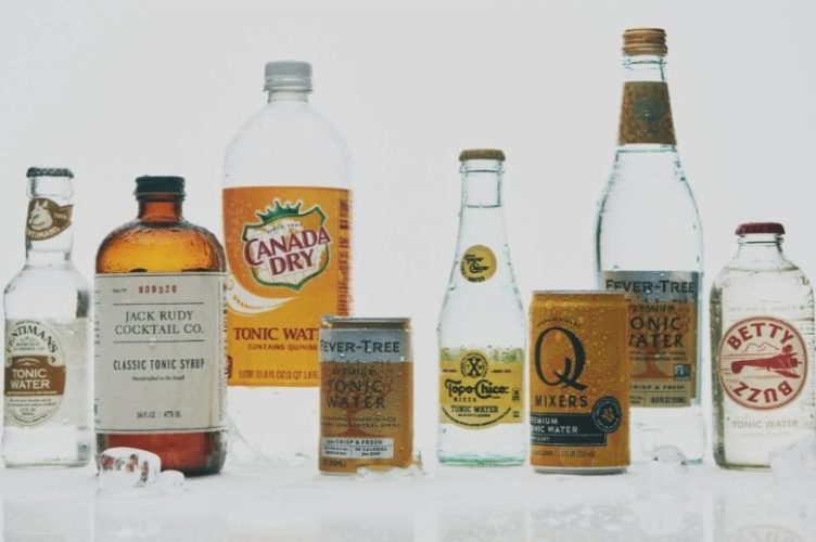 A chilled glass of gin and tonic with tonic water bottles and citrus garnishes arranged on a home bar.
