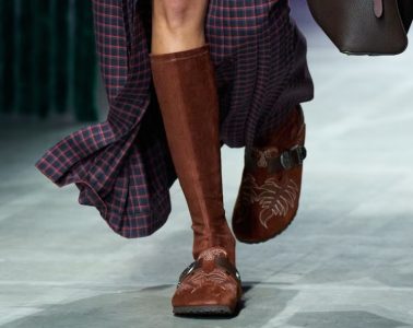 A pair of Birkenstock by Etro shoes showcases suede, paisley-inspired detailing, fringe, and Western-style hardware against an earthy backdrop.