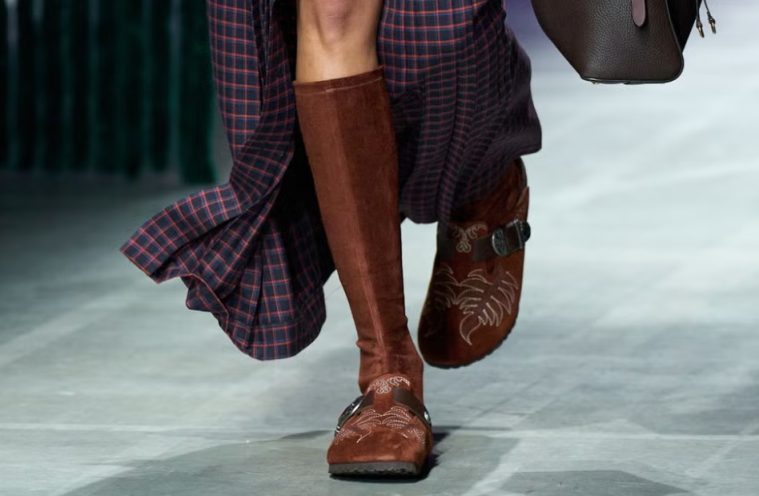 A pair of Birkenstock by Etro shoes showcases suede, paisley-inspired detailing, fringe, and Western-style hardware against an earthy backdrop.