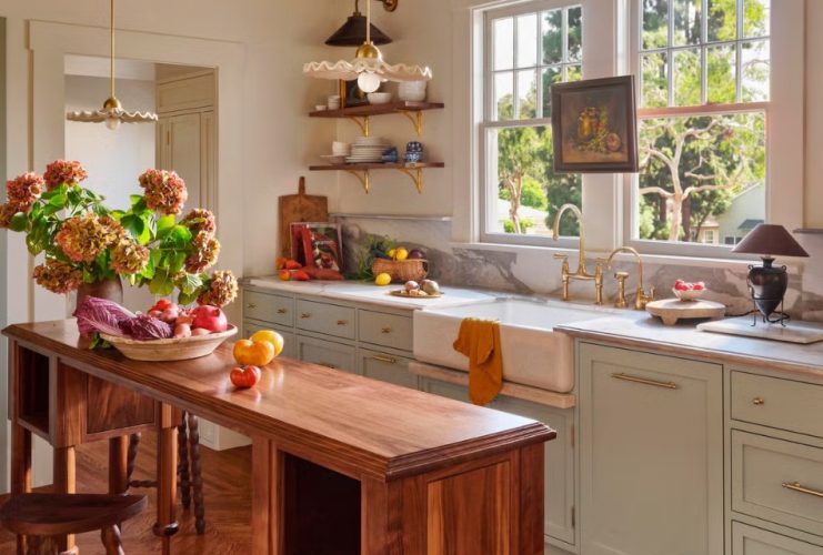 A bright 194-square-foot cottage kitchen features green cabinetry, warm wood, marble counters, exposed beams, and a central island filled with fresh produce.